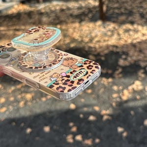 May include: A clear phone case with a leopard print design featuring a cartoon character and the word "Kitty." The case has a matching pop-up grip with a clear top filled with glitter. The phone case is on a blurred outdoor background.