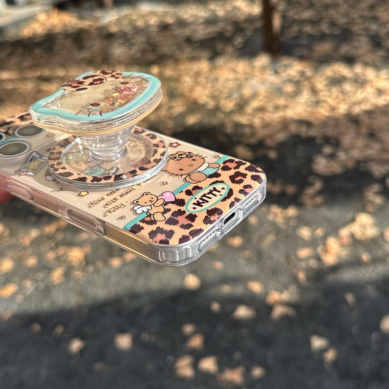 May include: A clear phone case with a leopard print design featuring a cartoon character and the word "Kitty." The case has a matching pop-up grip with a clear top filled with glitter. The phone case is on a blurred outdoor background.