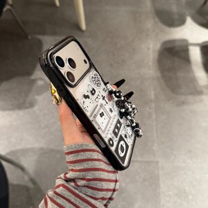 Op de afbeelding: Zwarte en transparante telefoonhoes met een decoratief ontwerp. De hoes heeft een zwarte rand en een transparante achterkant met een patroon van zwart-witte illustraties. Een 3D-versiering van zwarte en witte kralen en strikken is aan de zijkant bevestigd.