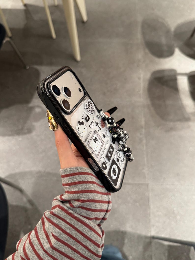 Op de afbeelding: Zwarte en transparante telefoonhoes met een decoratief ontwerp. De hoes heeft een zwarte rand en een transparante achterkant met een patroon van zwart-witte illustraties. Een 3D-versiering van zwarte en witte kralen en strikken is aan de zijkant bevestigd.