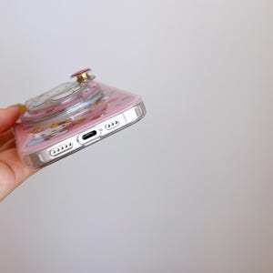 May include: Pink phone case with a clear, raised pop-up design featuring a cartoon character. The case has a silver border and a pink background with character illustrations. The pop-up has a gold-colored button.