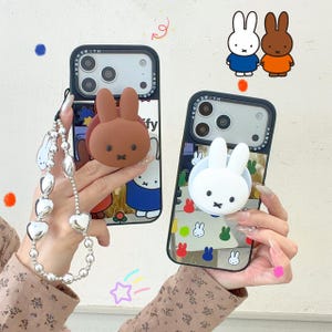 May include: Two black smartphone cases featuring Miffy character designs. One case has a brown rabbit grip and a silver heart-bead phone charm. The other case has a white rabbit grip and a mirrored back with small colorful rabbit icons printed on it.