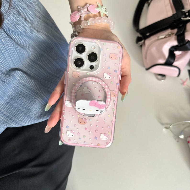 May include: A pink phone case with a clear ring holder featuring a Hello Kitty design. The case has a pink background with repeating Hello Kitty faces and stars. The ring holder has a white Hello Kitty face with a pink bow.