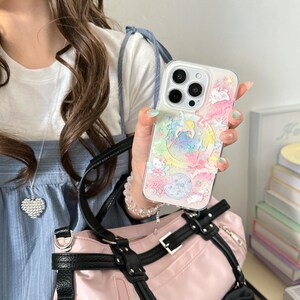 May include: Clear phone case with a colorful design featuring cartoon characters, a crescent moon, and stars. The case has a raised, textured design and is held in a person's hand. The case is on a pink bag.