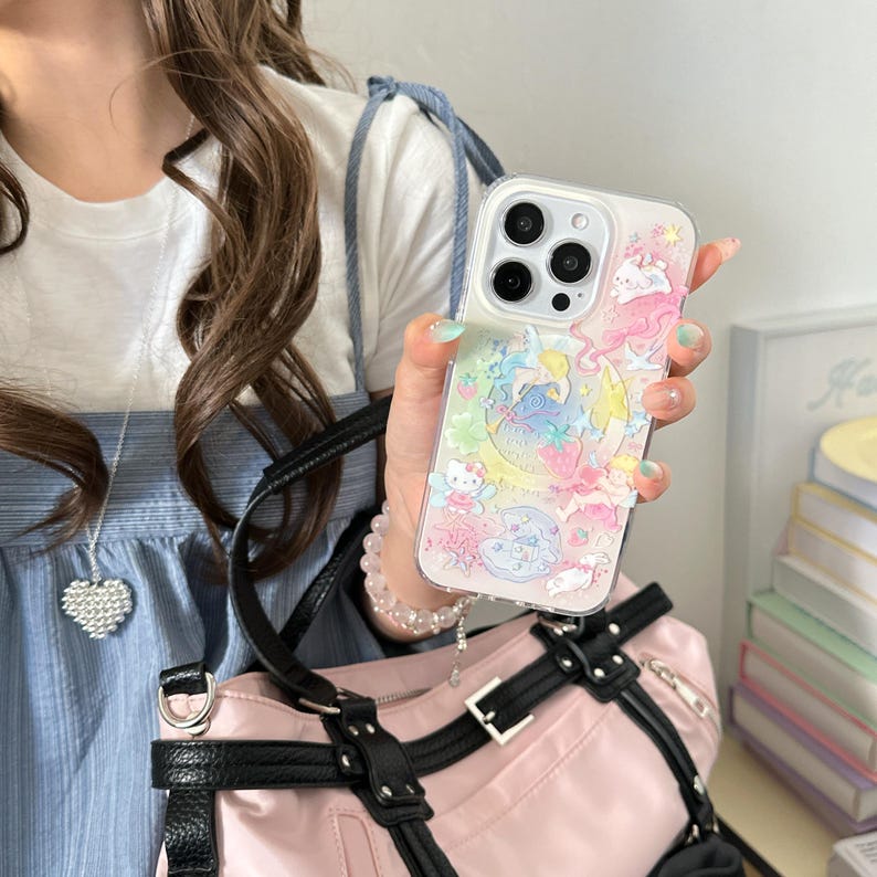May include: Clear phone case with a colorful design featuring cartoon characters, a crescent moon, and stars. The case has a raised, textured design and is held in a person's hand. The case is on a pink bag.