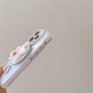 K&ouml;nnte beinhalten: Transparente Handyh&uuml;lle mit einer rosa-wei&szlig;en Schleife und einem Hello Kitty-Design. Die H&uuml;lle hat einen rosa Rand und einen Ringhalter. Der Kamerabereich hat einen rosa Ring um die Linsen.