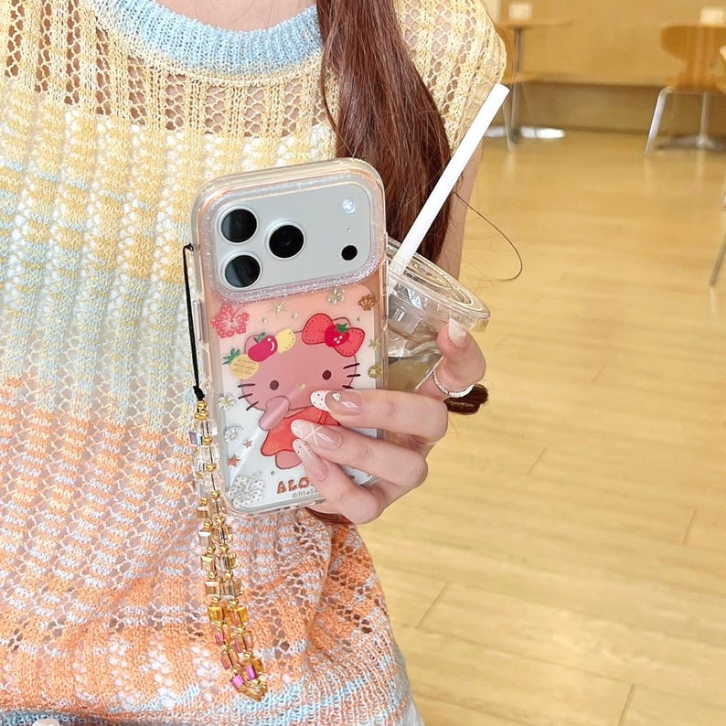 May include: Clear phone case featuring a Hello Kitty design with a red bow and strawberry accents. The case has a decorative strap with gold and pink beads. The phone case is designed to fit a smartphone.