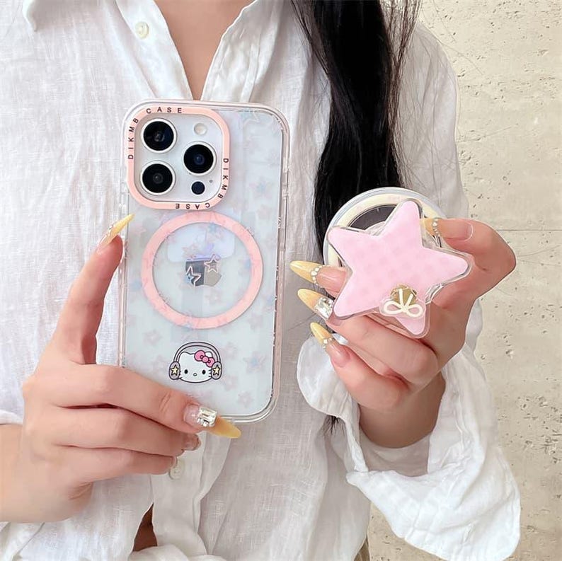 K&ouml;nnte beinhalten: Transparente Handyh&uuml;lle mit rosa Rand und Hello Kitty-Design. Die H&uuml;lle hat einen rosa Ring und einen sternf&ouml;rmigen Handyhalter. Der Halter ist rosa mit einem Schleifendesign. Die Handyh&uuml;lle ist mit kleinen Blumenformen verziert.