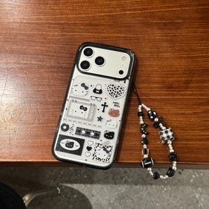 Op de afbeelding: Transparante telefoonhoes met een zwarte rand en diverse cartoonontwerpen, waaronder Hello Kitty-personages, harten en strikken. Het heeft een kralen polsbandje met zwarte en witte kralen.