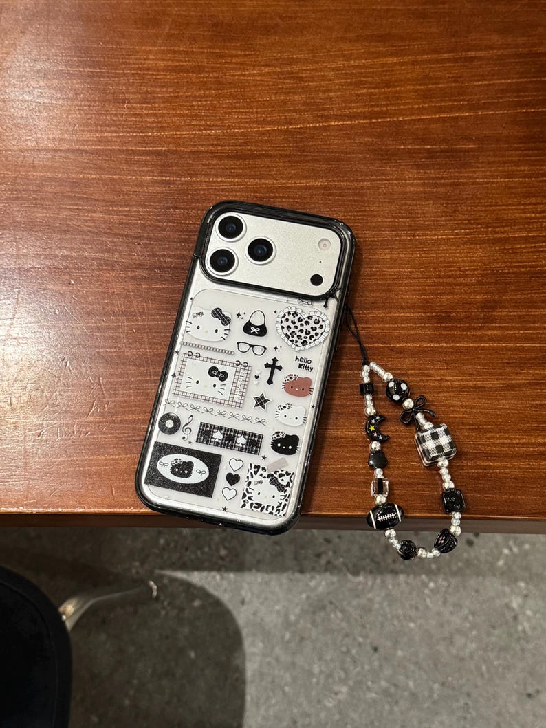 Op de afbeelding: Transparante telefoonhoes met een zwarte rand en diverse cartoonontwerpen, waaronder Hello Kitty-personages, harten en strikken. Het heeft een kralen polsbandje met zwarte en witte kralen.