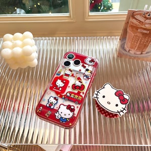 May include: A clear phone case with a red border, featuring Hello Kitty designs. The case has multiple images of Hello Kitty, a red bus, and stars. A Hello Kitty phone grip is also visible.