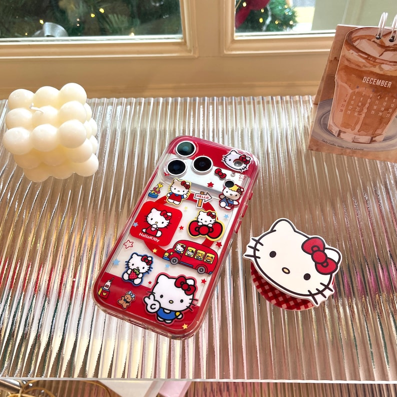 May include: A clear phone case with a red border, featuring Hello Kitty designs. The case has multiple images of Hello Kitty, a red bus, and stars. A Hello Kitty phone grip is also visible.