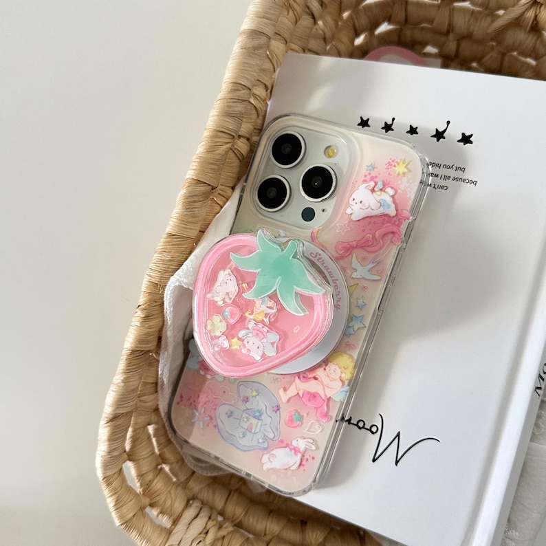 May include: Clear phone case with a pink strawberry pop socket. The case features a pink and white design with cartoon characters and the word "Strawberry". The phone case is inside a woven basket.