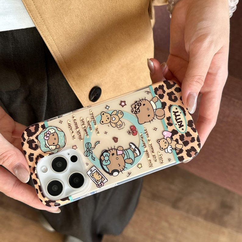 May include: Clear phone case with a leopard print border and a cartoon design featuring a cat and teddy bears. The case has text that reads "Kitty" and other phrases. The case is held by a person.
