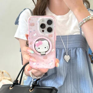 May include: Pink phone case featuring a Hello Kitty design. The case has a clear back with a pattern of Hello Kitty faces and stars. A circular pop-up grip with a Hello Kitty face is attached to the back. The case is designed for a smartphone.