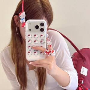 K&ouml;nnte beinhalten: Transparente Handyh&uuml;lle mit einem Muster aus Hello Kitty-Gesichtern. Die H&uuml;lle ist mit einem Anh&auml;nger mit einem roten Herz und einer Hello Kitty-Figur verziert. Eine rote Haarspange mit einer Hello Kitty-Figur ist ebenfalls sichtbar.