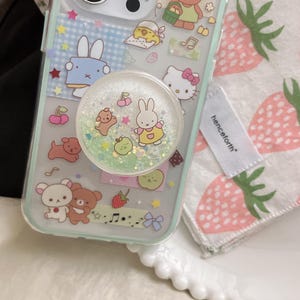 May include: A clear phone case decorated with cartoon characters, including a bunny, bear, and Hello Kitty. A glittery pop socket with a bunny design is attached. A white tag with the word "henceforth" is visible.