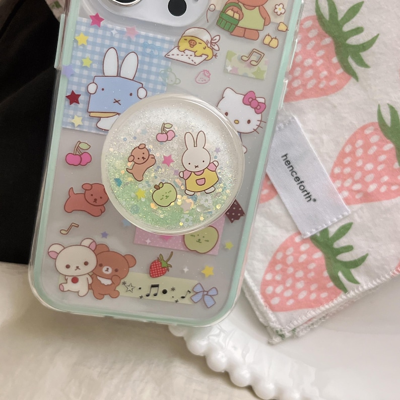 May include: A clear phone case decorated with cartoon characters, including a bunny, bear, and Hello Kitty. A glittery pop socket with a bunny design is attached. A white tag with the word "henceforth" is visible.