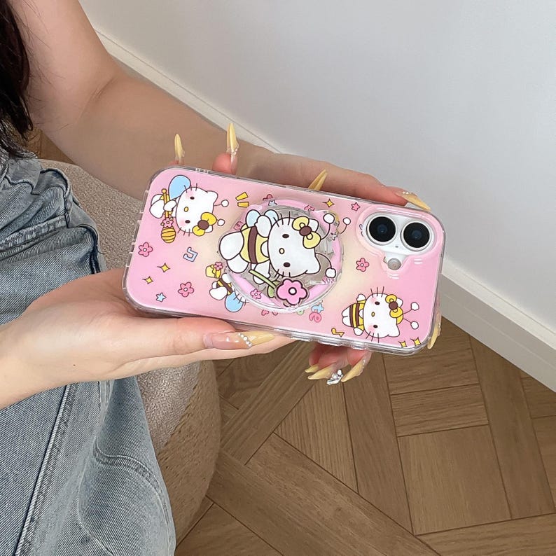 May include: Pink phone case featuring Hello Kitty and bee-themed designs. The case has a clear border and a pop-up grip. The design includes Hello Kitty characters dressed as bees, flowers, and other decorative elements.