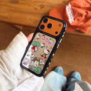 May include: Black and orange phone case with a clear window featuring Hello Kitty and other cartoon characters. The case has a black border with white accents and a clear back. The phone case is on a wooden surface.