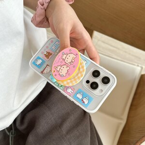 Puede incluir: Funda transparente para tel&eacute;fono con una variedad de dise&ntilde;os de pegatinas coloridas, incluyendo personajes de dibujos animados y un agarre emergente amarillo y rosa con Hello Kitty. La funda tiene un borde blanco y un recorte para c&aacute;mara de triple lente.