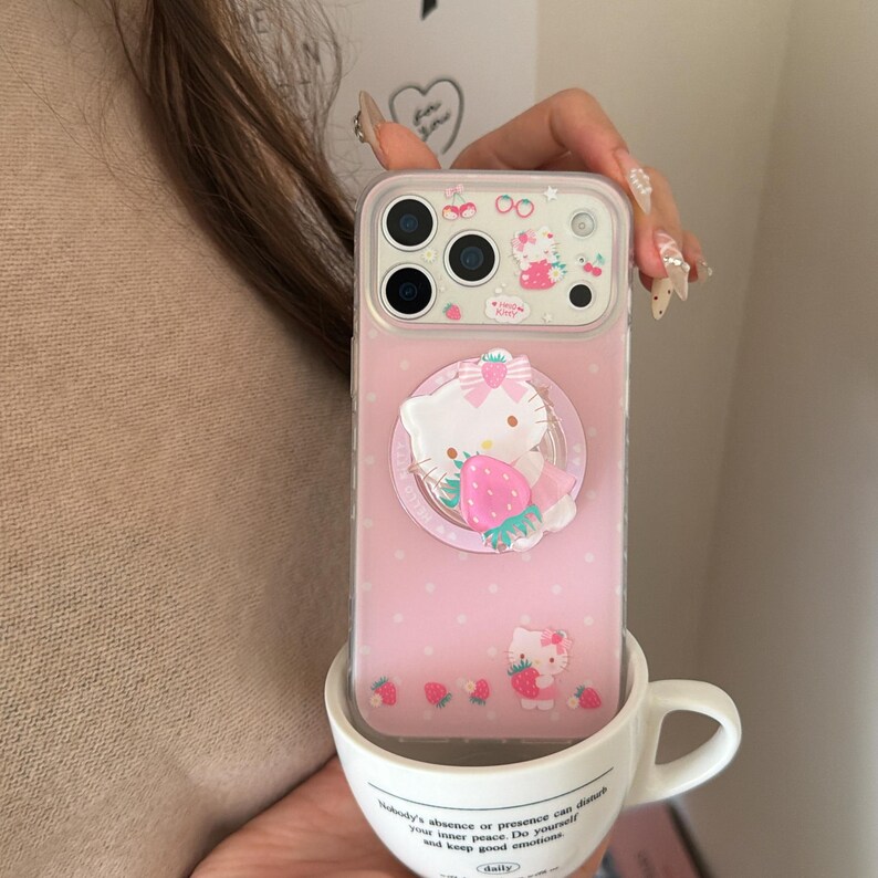 May include: A pink phone case featuring a Hello Kitty design with a strawberry. The case has a clear border and is decorated with small pink polka dots and strawberry illustrations. The phone case is in a white teacup.