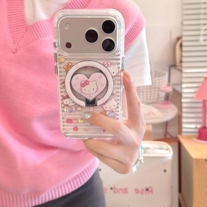 May include: Clear phone case featuring a Hello Kitty design with a heart-shaped ring holder. The case has a pink and white striped pattern with illustrations of Hello Kitty and other decorative elements.