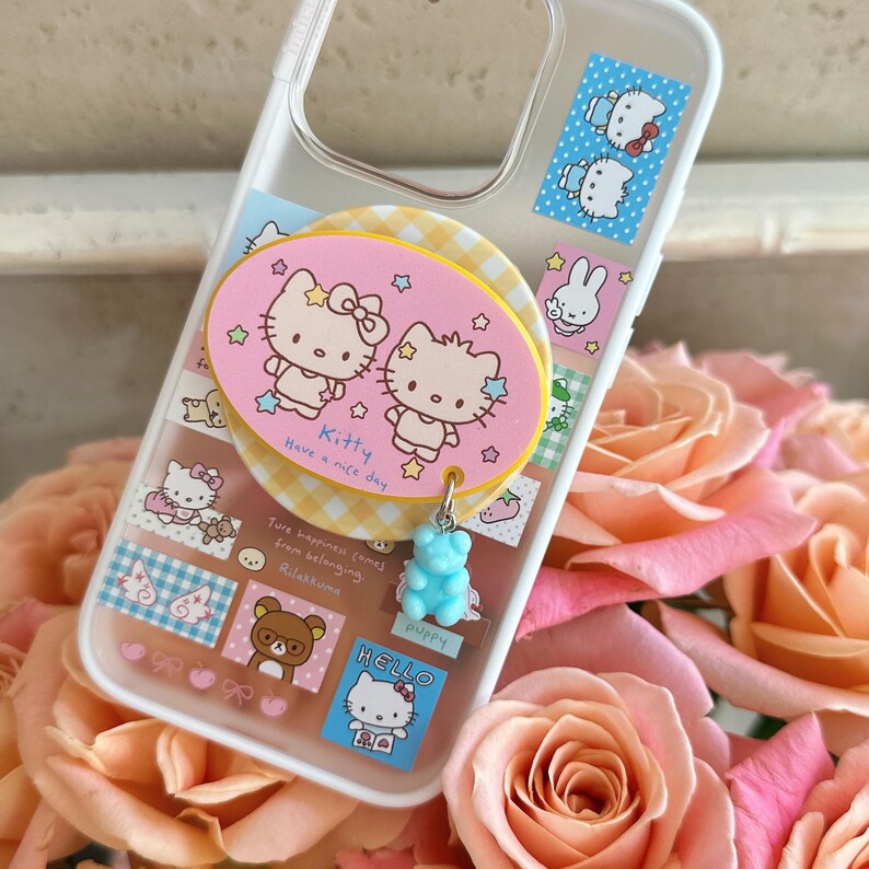 Puede incluir: Funda transparente para tel&eacute;fono con un dise&ntilde;o rosa y amarillo con personajes de dibujos animados. La funda tiene un charm de un osito de goma azul. El texto "Kitty Have a nice day" es visible.