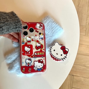 May include: Red phone case with Hello Kitty graphics, including the character in various poses, a bus, and stars. A matching white and red pop socket with the Hello Kitty face is also visible. The case is on a white surface with a blue fuzzy background.