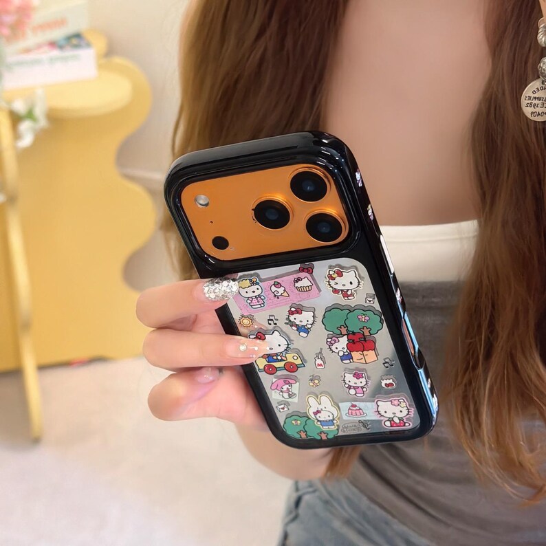 May include: Black and gold phone case with a clear panel featuring Hello Kitty and other cartoon characters. The case has a glossy finish and a protective bumper around the edges. The design includes various colorful illustrations of the popular character.