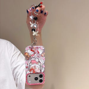 May include: Clear phone case with a pink border featuring Hello Kitty characters. The case is attached to a beaded lanyard with white bows, black and silver beads, and Hello Kitty charms.