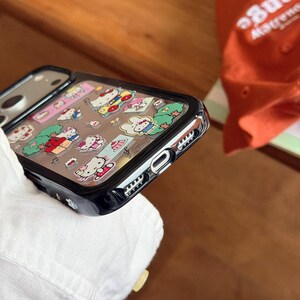 May include: A black phone case with a clear back featuring colorful Hello Kitty character designs. The case has a black border and a clear, protective layer over the images. The phone case is shown at an angle.