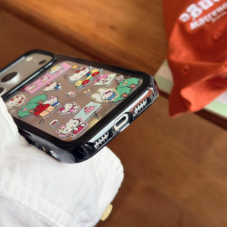 May include: A black phone case with a clear back featuring colorful Hello Kitty character designs. The case has a black border and a clear, protective layer over the images. The phone case is shown at an angle.