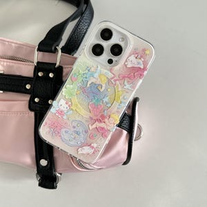 May include: Clear phone case with a colorful, whimsical design featuring cherubs, stars, a crescent moon, and strawberries. The case is resting on a pink handbag with black straps.