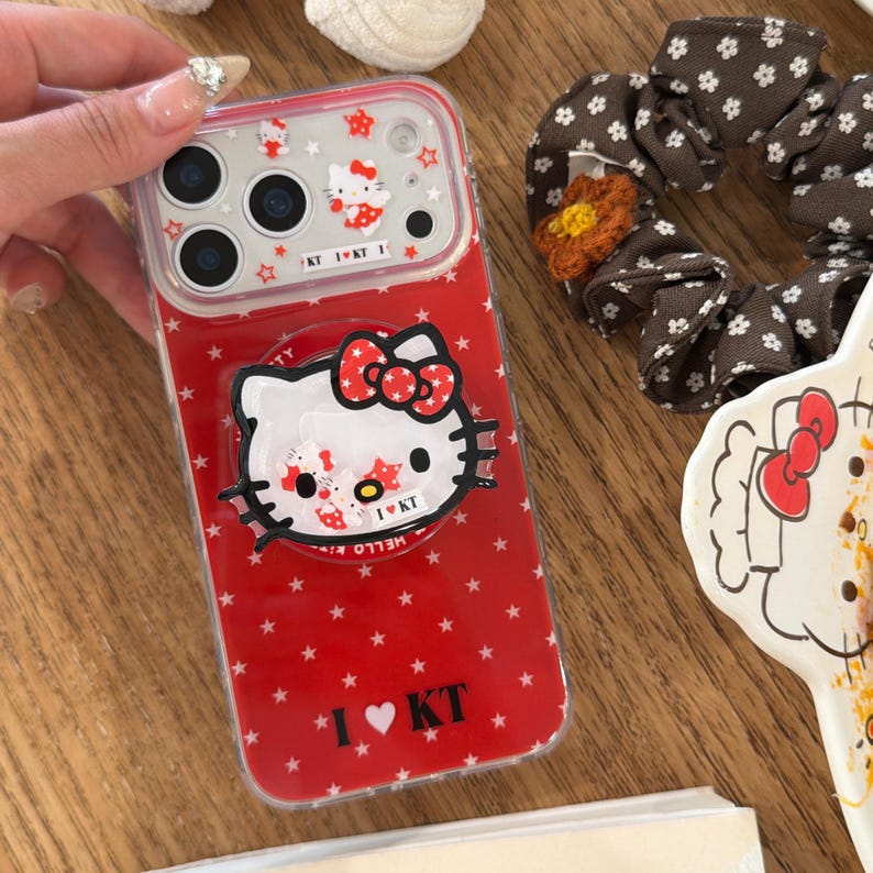 May include: Red phone case featuring a Hello Kitty design with a large bow and star accents. The case has a clear border and includes the text "I ❤️ KT". The case is designed for a smartphone.
