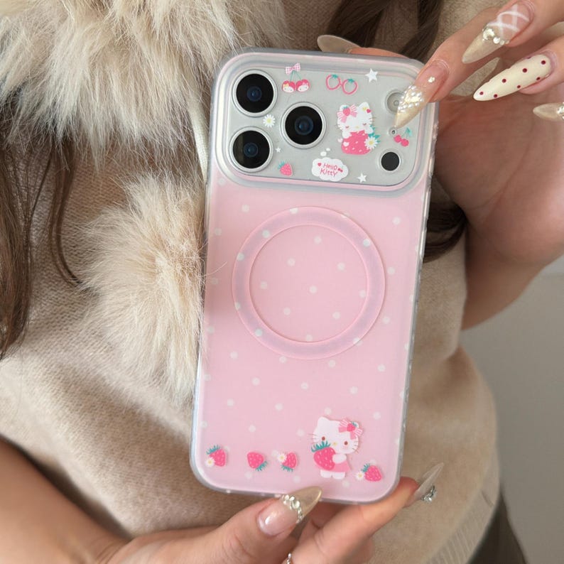 May include: Pink phone case with a Hello Kitty design. The case features a clear border, a pink back with white polka dots, and a circular design. Hello Kitty is depicted with strawberries. The case is designed for a smartphone.