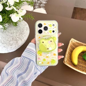 Könnte beinhalten: Transparente Handyhülle mit hellgrünem Rand und Frosch-Motiv. Die Hülle verfügt über einen Pop-up-Froschgriff und ist mit verschiedenen Frosch-Illustrationen in Grün- und Gelbtönen verziert. Für Smartphones.