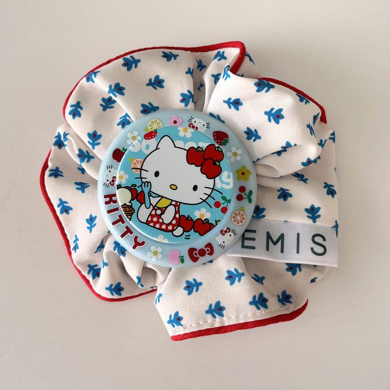 May include: A white scrunchie with a blue floral pattern and red trim. A light blue circular pin featuring Hello Kitty with cherries and the word "KITTY" is attached. The tag reads "EMIS".