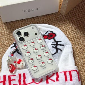 K&ouml;nnte beinhalten: Transparente Handyh&uuml;lle mit einem Muster von Hello Kitty-Gesichtern, die verschiedene Emotionen ausdr&uuml;cken. Die H&uuml;lle liegt auf einer wei&szlig;en M&uuml;tze mit rotem Text und einer Hello Kitty-Grafik. Ein Anh&auml;nger mit roten Herz- und Hello Kitty-Akzenten h&auml;ngt am Telefon.