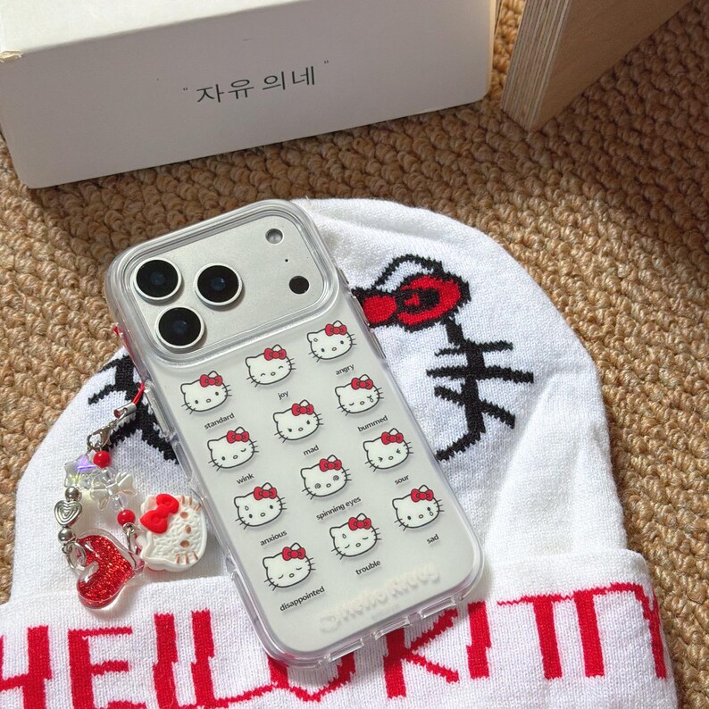 K&ouml;nnte beinhalten: Transparente Handyh&uuml;lle mit einem Muster von Hello Kitty-Gesichtern, die verschiedene Emotionen ausdr&uuml;cken. Die H&uuml;lle liegt auf einer wei&szlig;en M&uuml;tze mit rotem Text und einer Hello Kitty-Grafik. Ein Anh&auml;nger mit roten Herz- und Hello Kitty-Akzenten h&auml;ngt am Telefon.