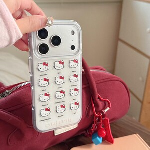 K&ouml;nnte beinhalten: Transparente Handyh&uuml;lle mit einem sich wiederholenden Muster von Hello Kitty-Gesichtern, die verschiedene Emotionen zeigen. Die H&uuml;lle hat einen transparenten Rand und eine wei&szlig;e Basis mit dem Text "(c) Sanrio Co., Ltd.".