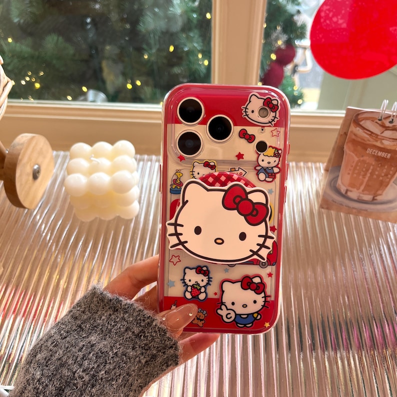 May include: A red and clear phone case featuring Hello Kitty designs. The case has a red border and a clear back, showcasing multiple Hello Kitty illustrations with bows and stars. The case is held by a person.