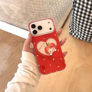 Puede incluir: Funda de tel&eacute;fono roja con un dise&ntilde;o de manzana de Hello Kitty dentro de un recorte en forma de coraz&oacute;n. La funda tiene un borde transparente y est&aacute; decorada con peque&ntilde;as estrellas doradas. La funda del tel&eacute;fono se sostiene en una mano.
