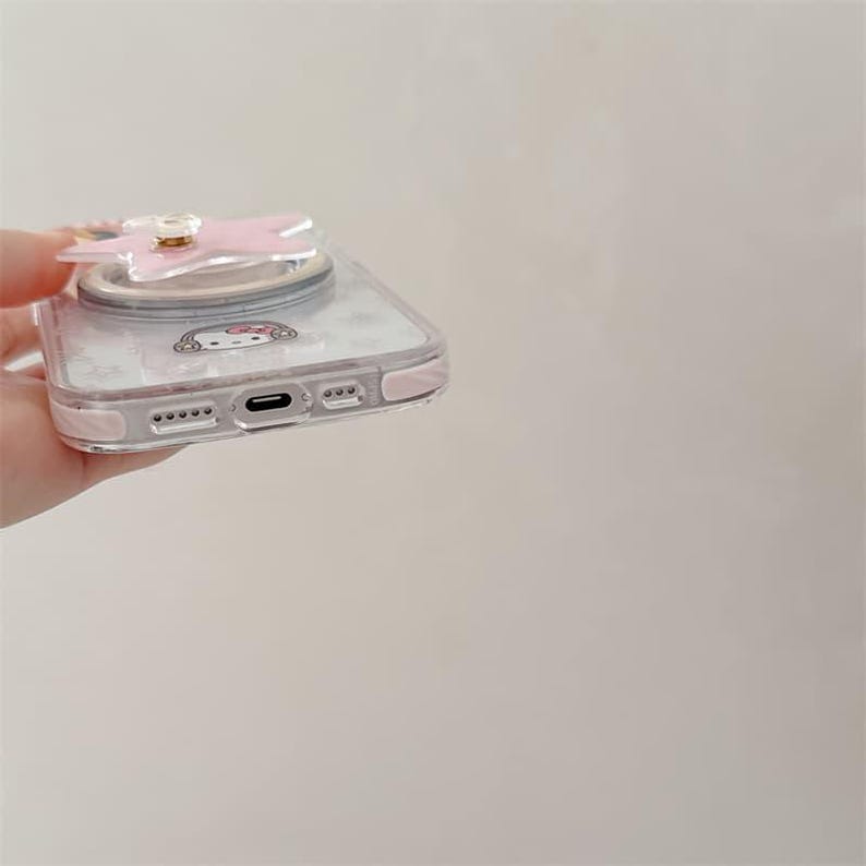 K&ouml;nnte beinhalten: Transparente Handyh&uuml;lle mit einem rosa schleifenf&ouml;rmigen Handyhalter. Die H&uuml;lle hat einen rosa Rand und ein Cartoon-Charakter-Design. Die H&uuml;lle ist f&uuml;r ein Smartphone konzipiert.
