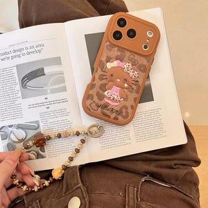Läderfodral i rosa brunt leopardmönster med Hello Kitty-motiv och snodd för iPhone 17 Pro, 16 Pro Max och 15-serien. Phone case + strap