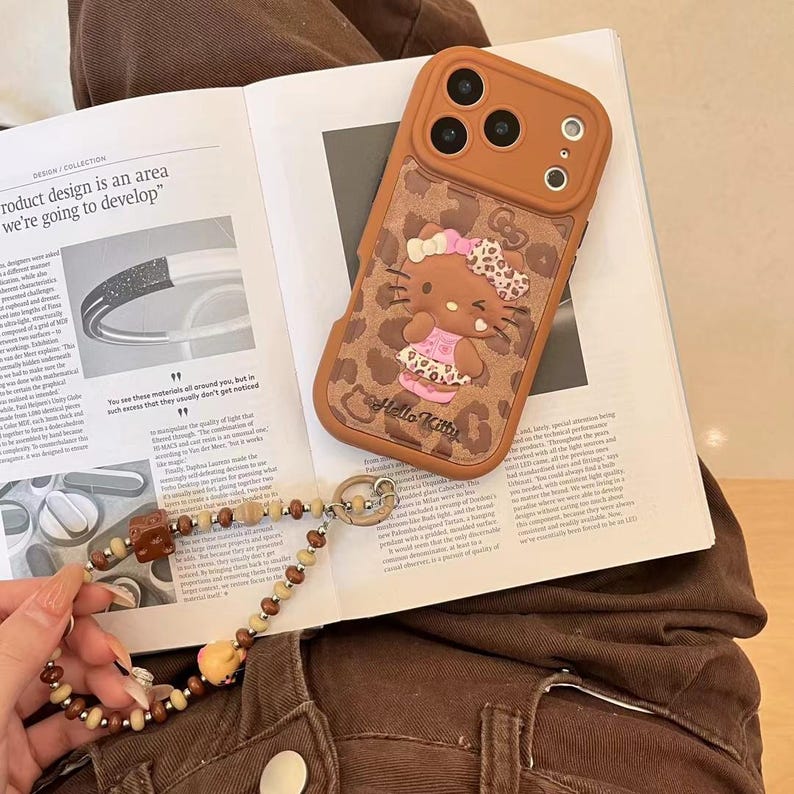 Läderfodral i rosa brunt leopardmönster med Hello Kitty-motiv och snodd för iPhone 17 Pro, 16 Pro Max och 15-serien. Phone case + strap