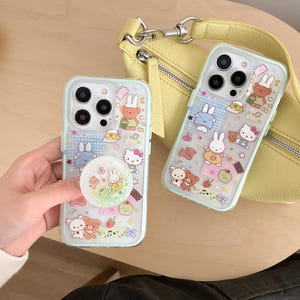 May include: Two clear phone cases with cartoon character designs. One case is held by a hand. The cases feature a light blue border and a pop socket. A yellow purse is in the background.