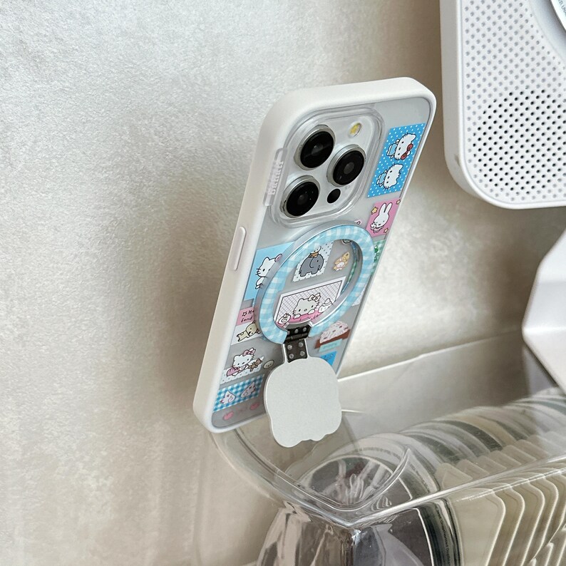 Puede incluir: Funda de tel&eacute;fono blanca con parte trasera transparente con un dise&ntilde;o de dibujos animados colorido. La funda tiene un soporte de anillo incorporado con un patr&oacute;n de cuadros azul. La funda del tel&eacute;fono est&aacute; apoyada en un soporte de pl&aacute;stico transparente.