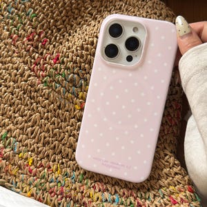 May include: A pale pink phone case adorned with white polka dots. The case features a camera aperture and the inscription "seize the moment of happiness". The phone case rests on a woven straw bag.