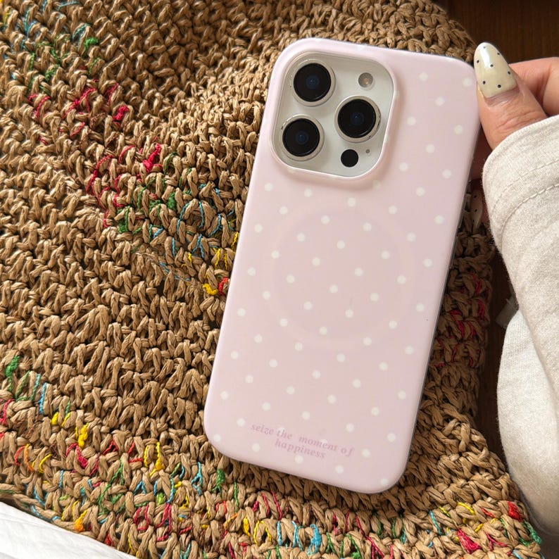 May include: A pale pink phone case adorned with white polka dots. The case features a camera aperture and the inscription "seize the moment of happiness". The phone case rests on a woven straw bag.